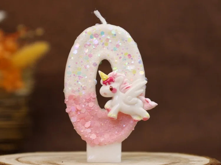 Una vela número cero blanca decorada con purpurina rosa y una pequeña figura de unicornio blanco.