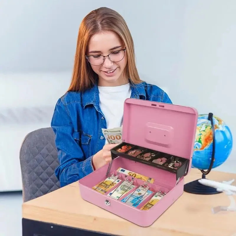 A young woman is holding a pink lockable metal cash box with an organizer tray and two keys on a wooden table.