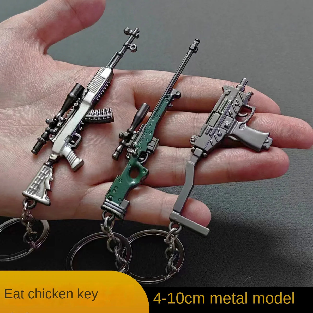 A hand holds a realistic miniature gun model keychain featuring an assault rifle and a pistol, made of metal.