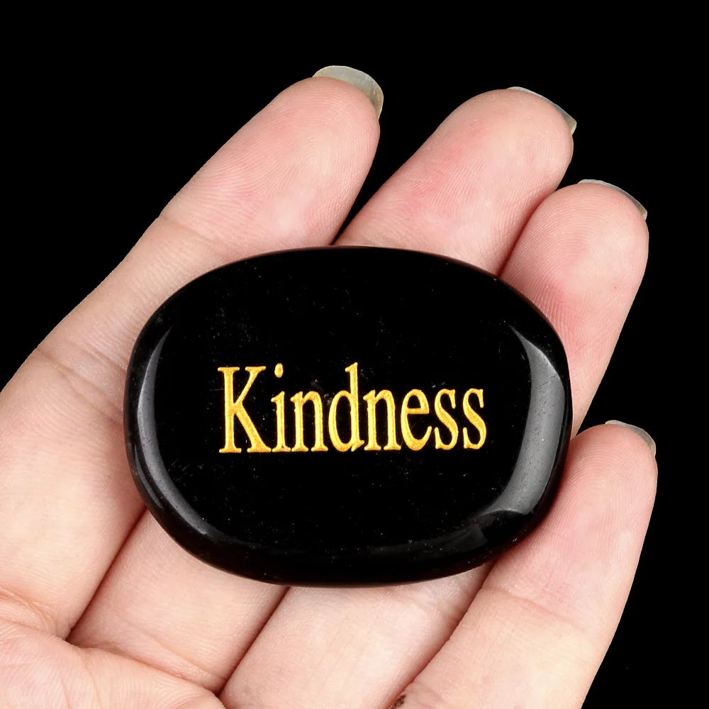 A black oval natural crystal with the word Kindness engraved on it is held in a hand.