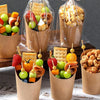 Several disposable kraft paper serving cups filled with various fruit and nut snacks like apples, strawberries, and granola.