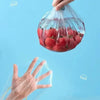 A hand is holding a clear plastic food wrap cover filled with red tomatoes against a light blue background.