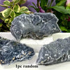 Three irregular dark gray natural zinc crystal clusters displayed on a white surface with green foliage in the background