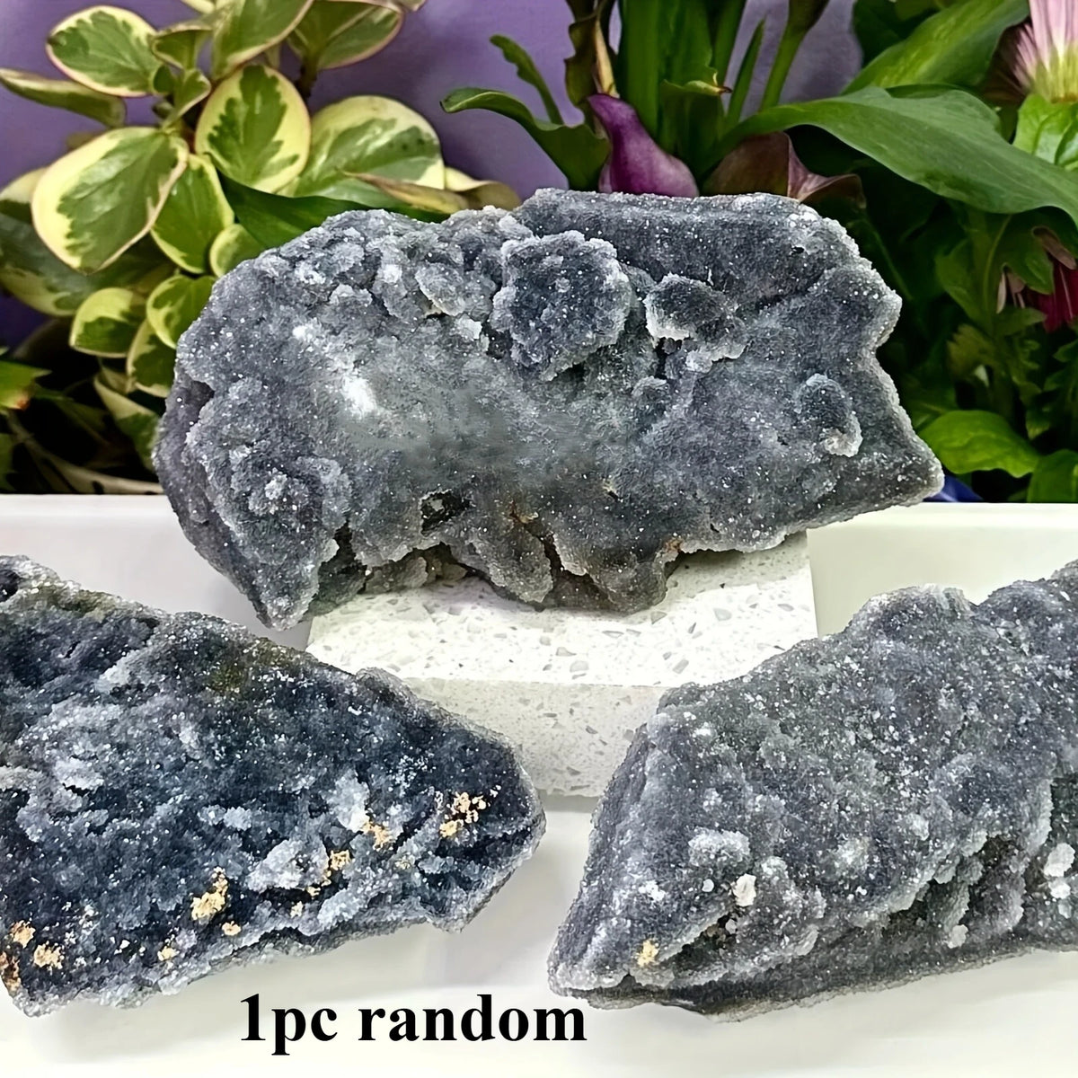 Three irregular dark gray natural zinc crystal clusters displayed on a white surface with green foliage in the background