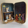 A tin box containing a nativity scene decoration featuring hand-painted figures, stars, and a starry background.