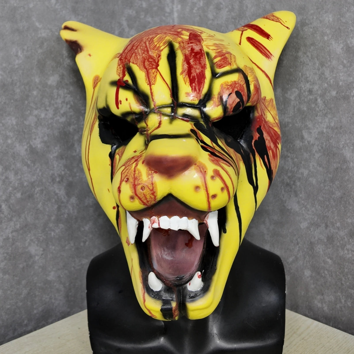 A yellow tiger horror latex mask with red blood splatters and black markings, displayed on a black bust.