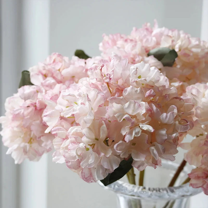 A close-up of a pink silk hydrangea stem with vintage burnt edge petals in a clear glass vase.
