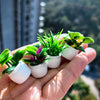 A hand holds a set of miniature potted plants with vibrant green and purple leaves for dollhouse decor.