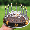 A dark chocolate birthday cake decorated with lit candles and star toppers, held in a hand against a green outdoor background