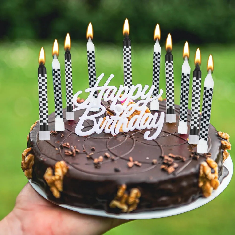 A dark chocolate birthday cake decorated with lit candles and star toppers, held in a hand against a green outdoor background