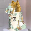 A tiered cake decorated with light green and gold accents, featuring palm leaf toppers and floral elements.