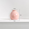 A pink ceramic mini vase with a rounded body and narrow neck, shown on a white shelf.