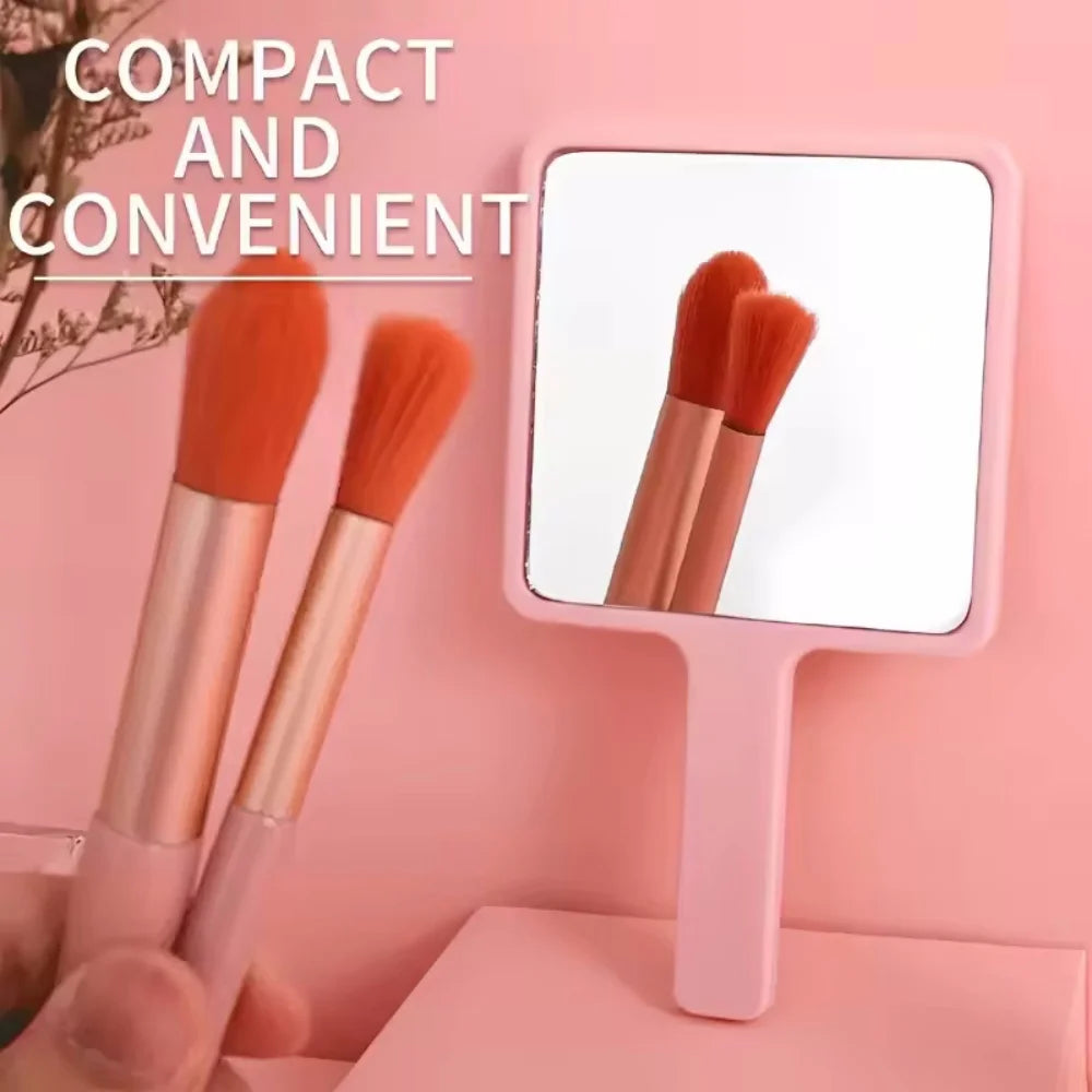 A pink compact handheld makeup mirror shows two orange-tipped makeup brushes.