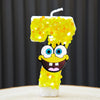 A yellow sponge cake candle shaped like the number 7, decorated with gold glitter and a SpongeBob face.