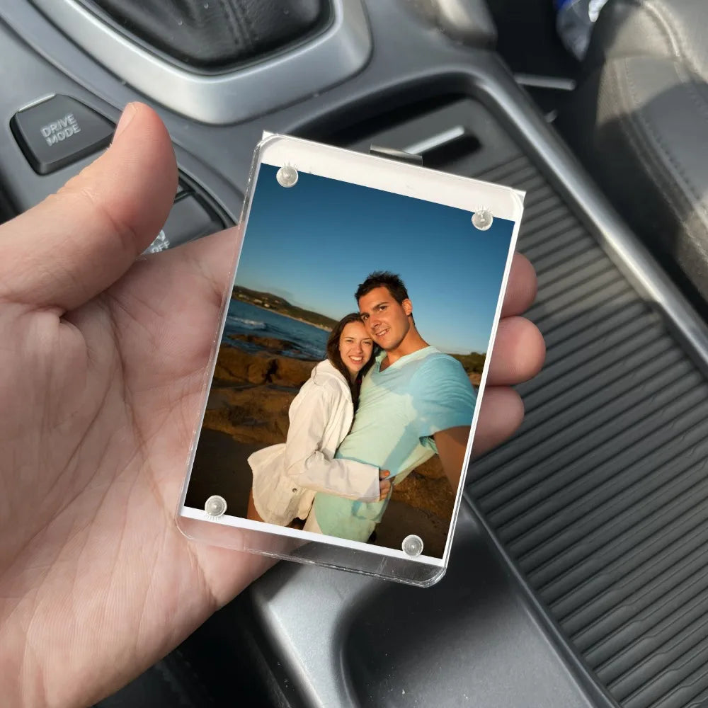 A photo frame displaying a couple smiling outdoors, held by a hand against a car interior.