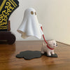 A white ghost figurine is shown hovering above a small dog figurine on a wooden desk.