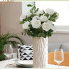 A bouquet of white artificial roses in a textured vase sits on a white table next to glassware and greenery.