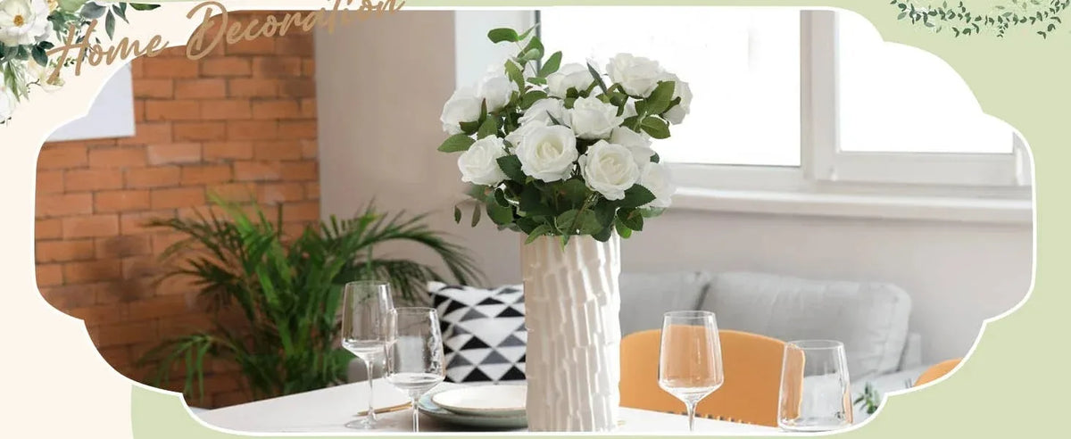 A bouquet of white artificial roses in a textured vase sits on a white table next to glassware and greenery.