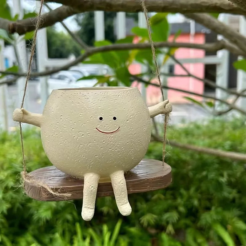 A light beige resin ornament shaped like a smiling face is suspended by twine against a blurred green outdoor background.