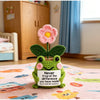 Hand crocheted frog ornament with a pink flower growing from its back on a pink rug