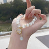 A hand is holding a bracelet made of small, round white natural stones with a gold charm.