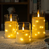 Three glass cylindrical candles with glowing LED star string lights on a woven fabric surface.