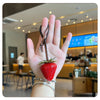 A hand holds a red strawberry charm and keychain against a blurred background of an indoor cafe.