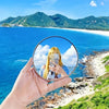 A hand holds a round convex mirror reflecting a scenic tropical landscape with blue ocean and white sand beach.
