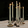 A set of six white taper candles in glass holders with lit flames, resting on a white cloth with pinecones and wood slices.