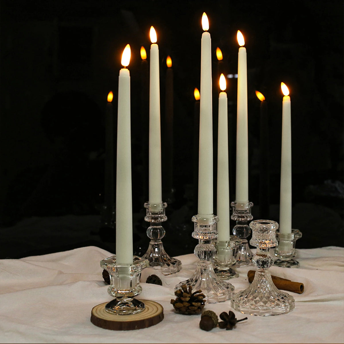 A set of six white taper candles in glass holders with lit flames, resting on a white cloth with pinecones and wood slices.