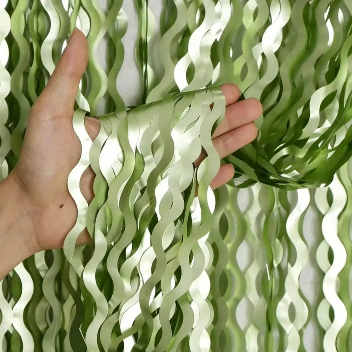 Sage green wavy foil fringe curtain streamers being held by a hand