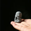 A small, gray stone statue of a monk with hands pressed to his face is held in an open palm against a dark background.