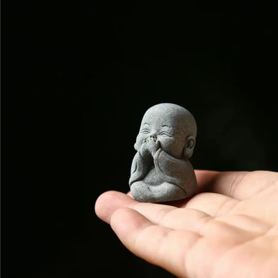 A small, gray stone statue of a monk with hands pressed to his face is held in an open palm against a dark background.