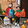Two children dressed in Halloween costumes holding orange treat bags with pumpkin and ghost designs in front of a brick wall.