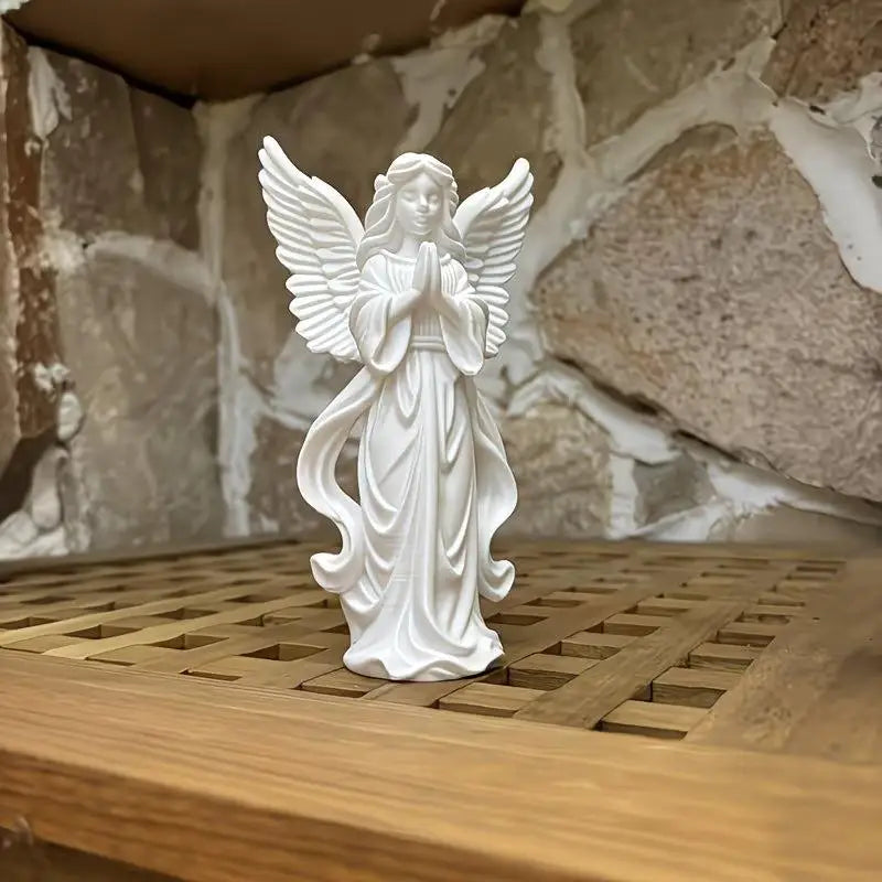 A white statue of a praying angel with wings stands on a wooden surface against a rustic stone wall background.