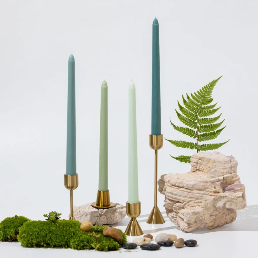 A collection of tall, gradient color taper candles in sage green and pale green set against a natural stone and moss backgrou