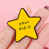 A hand holds a yellow star enamel pin with the text your did it on it.