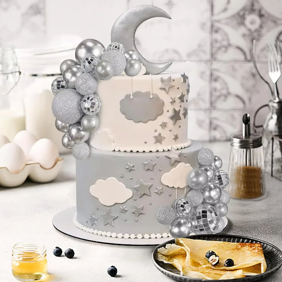 A tiered cake decorated with silver and gray disco ball-like toppers and clouds on a white background.
