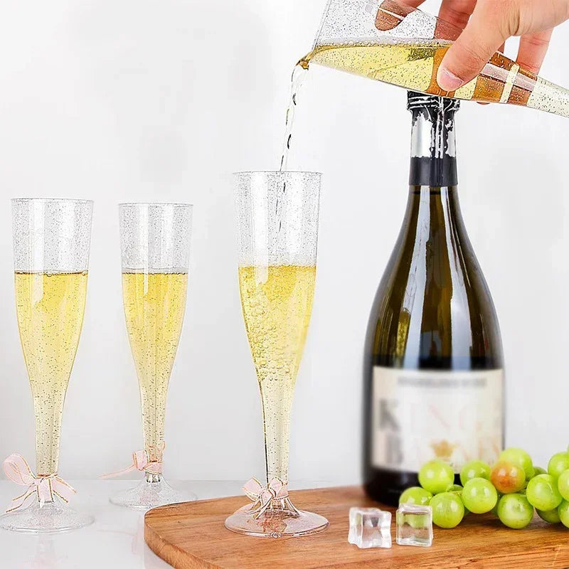 A hand pours sparkling yellow liquid into a glass, with two empty champagne flutes and green grapes visible.