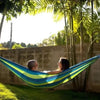 A person relaxing in a blue and green canvas camping hammock strung between two trees in a sunny backyard.