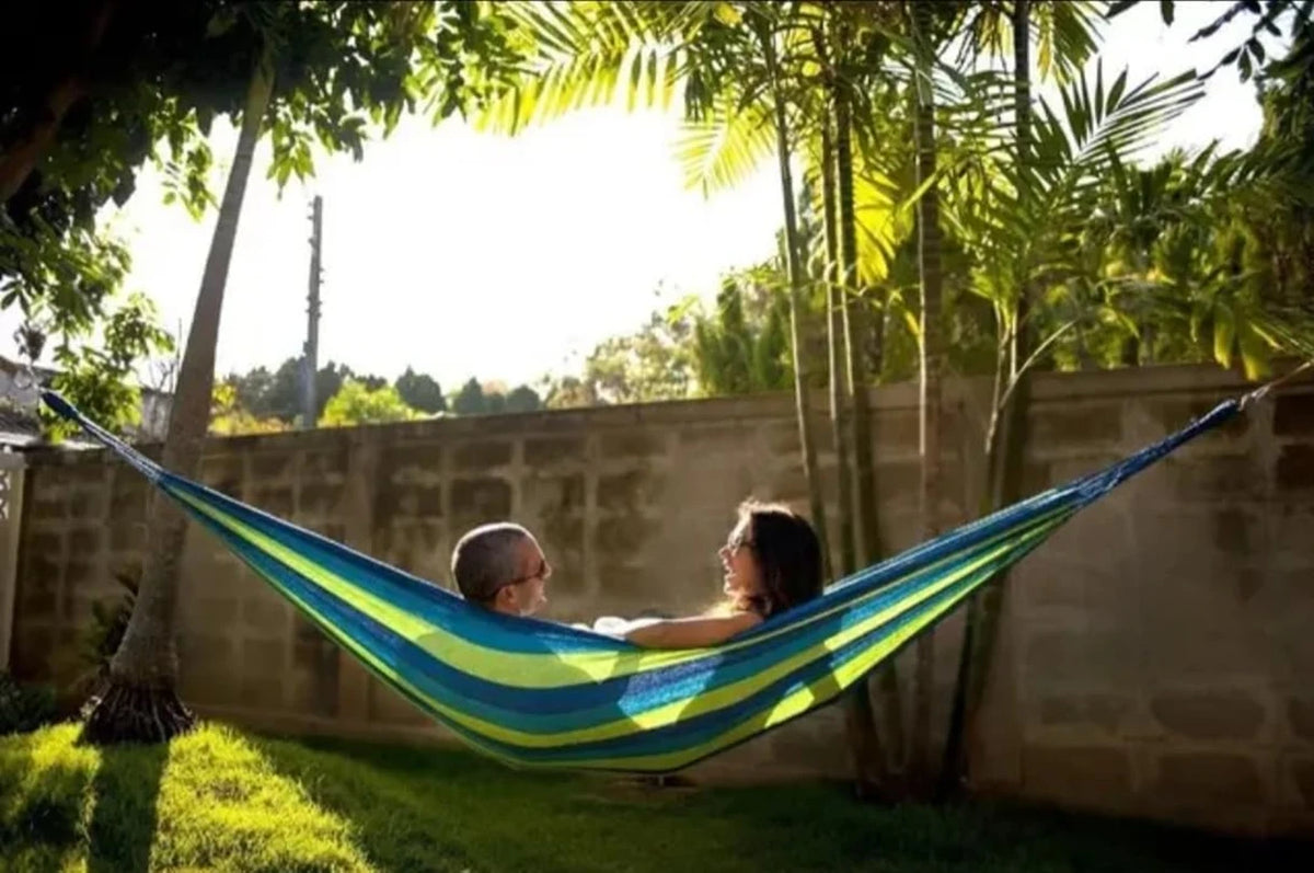 A person relaxing in a blue and green canvas camping hammock strung between two trees in a sunny backyard.