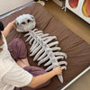 A person is posing with a white 3D dinosaur skeleton plush costume for kids on a brown blanket.