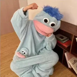 A person wearing a light blue monster onesie pajama with pink accents and blue hair, sitting down.