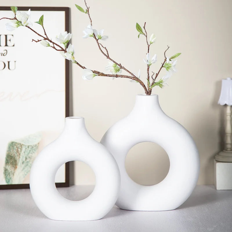 A white ceramic vase with a ring opening holds a branch of white blossoms on a light gray surface.