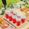 A tray of red fruit drinks in clear plastic cups served on a wooden table with crackers and grapes.