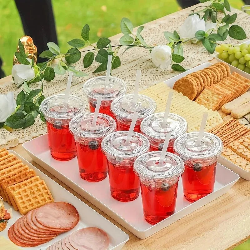 A tray of red fruit drinks in clear plastic cups served on a wooden table with crackers and grapes.