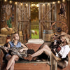 A woman sits on a wooden surface in front of a rustic barn door backdrop with a horse and western decor.