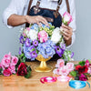 A person holds a bouquet of pink and purple flowers, including roses and hydrangeas, over a wooden table.