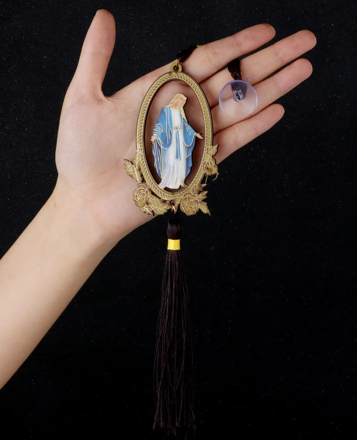 A hand holds a wooden pendant ornament depicting Our Lady of Grace with a blue and white robe.