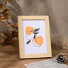 A light wooden photo frame with an orange abstract design is displayed on a wooden surface next to dried foliage.